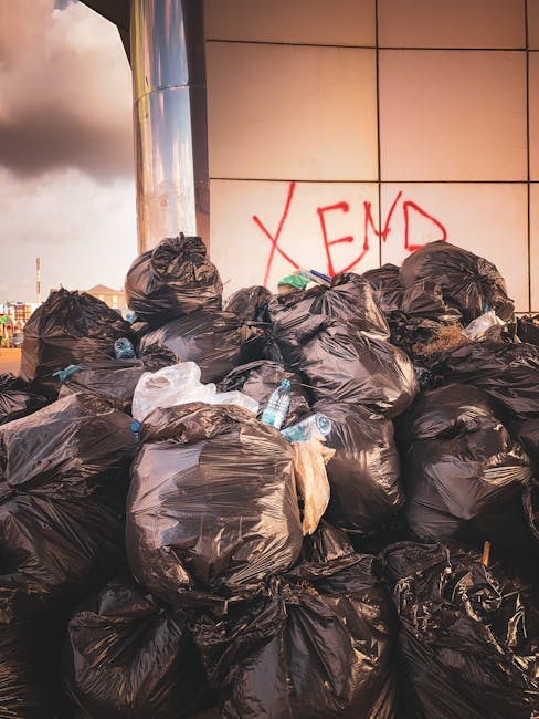 A large pile of black plastic rubbish bags filled with waste sits against a tiled wall outdoors, with some bags torn open revealing discarded plastic bottles and paper waste inside. The bags are stacked haphazardly, with a mix of tightly knotted and loosely closed sacks. Behind the pile, a graffiti marking with the word 'XEND' is spray-painted in red on the wall. In the background, an urban environment is faintly visible under a cloudy sky, suggesting the area is a site for unregulated waste disposal or a temporary rubbish collection point. The scene highlights issues related to waste management and the importance of proper rubbish removal services such as those offered by Rubbish Removal Balham, which specialise in alternative waste handling and on-site clearance solutions for private or commercial clients.