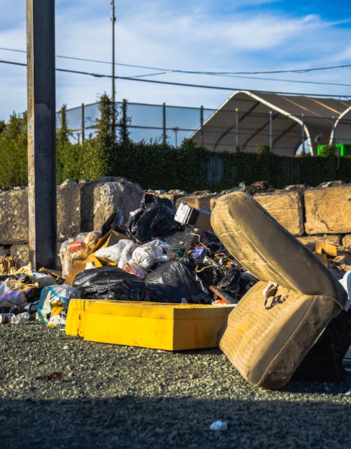 A pile of mixed rubbish is seen on the ground in an outdoor area, including black plastic bags filled with waste, a yellow plastic container, and a large, weathered car tyre that is leaning against a stone barrier. The debris appears to consist of household waste and discarded materials, with some items partially crushed or collapsed. In the background, a stone wall and a chain-link fence are visible, along with lush green shrubbery and trees. Further behind, there is a large structure with a curved, white roof, possibly a sports or exhibition hall, set against a partly cloudy sky with blue patches. The scene suggests a location where an independent rubbish removal service, such as Rubbish Removal Balham, might collect and clear bulky waste that is prepared for disposal or recycling, indicating an alternative waste handling scenario to council curbside collection. The environment appears to be a designated waste pickup or clearance point, emphasizing the need for professional rubbish removal services for efficient and safe waste management.