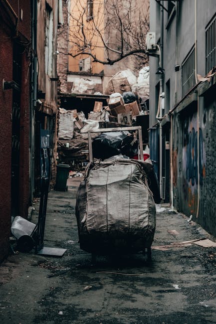 A narrow urban alleyway filled with an assortment of discarded rubbish and debris, including a large, grey, weathered bag in the foreground that appears to be made of a thick, fabric-like material, with visible tension lines and slight stains. Behind the bag, there are scattered smaller items such as plastic bottles, metal containers, and scraps of paper on the uneven, dark asphalt ground which shows signs of wear and oil stains. The alley is flanked by tall buildings on either side, with the left side featuring a rusty metal door and graffiti markings, while the right side has a grey wall with small barred windows and an external electrical box. In the background, a pile of assorted waste, including cardboard boxes and plastic bags, is stacked against a wooden fence, partially obscured by leafless tree branches overhead. The scene is lit by natural daylight, giving a dull atmosphere consistent with a private waste collection or clearance situation, typical of independent rubbish removal services. The overall environment emphasizes a space used for dumping refuse awaiting removal, typical of the type of rubbish removal service offered by Rubbish Removal Balham in the context of alternative waste handling efforts within urban settings.