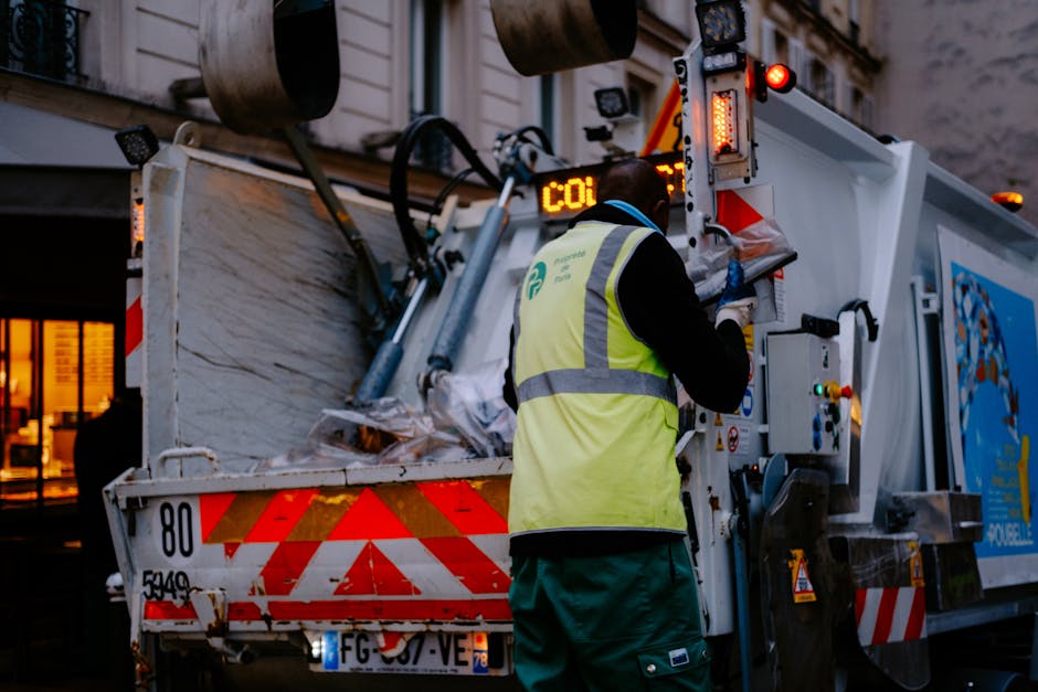 A uniformed waste collection worker wearing a high-visibility yellow vest and dark clothing operates a waste management vehicle, which features red and white reflective markings on the rear, in an urban setting during dusk. The vehicle's open back reveals a load of mixed rubbish, including crumpled paper, plastic, and packaging materials, while the worker interacts with control panels on the side of the truck. Background elements include a building with warmly lit windows and the street environment, emphasizing a typical street scene focused on rubbish collection and private waste handling services. The lighting suggests evening hours, with illuminated signals and ambient street lighting contributing to the scene's visibility, aligning with independent rubbish removal operations in the area.