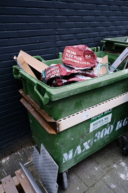 A uniformed waste collection worker wearing a high-visibility yellow vest and dark clothing operates a waste management vehicle, which features red and white reflective markings on the rear, in an urban setting during dusk. The vehicle's open back reveals a load of mixed rubbish, including crumpled paper, plastic, and packaging materials, while the worker interacts with control panels on the side of the truck. Background elements include a building with warmly lit windows and the street environment, emphasizing a typical street scene focused on rubbish collection and private waste handling services. The lighting suggests evening hours, with illuminated signals and ambient street lighting contributing to the scene's visibility, aligning with independent rubbish removal operations in the area.