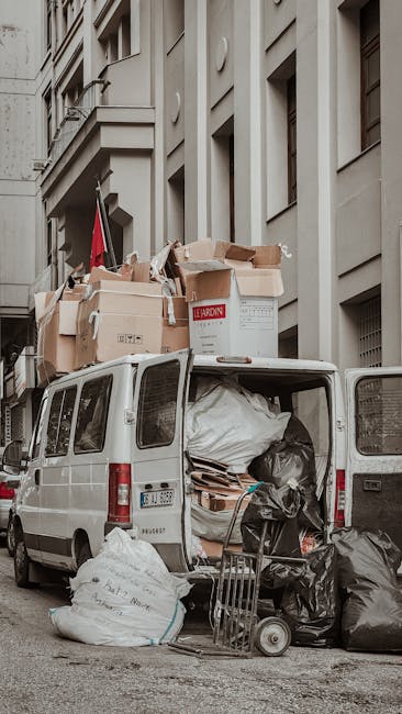 A white cargo van used for rubbish removal parked on a city street in front of a multi-storey building with a light grey facade featuring large vertical and horizontal architectural details. The van's open rear doors reveal a variety of waste materials, including black trash bags, cardboard boxes, and wooden pallets, closely packed inside and some spilling onto the pavement. Several cardboard boxes, some with printed labels, are stacked on top of the van, indicating on-site clearance or private waste collection activities typical of independent rubbish removal services. A hand truck loaded with additional black rubbish bags is positioned next to the open doors. The environment appears to be a typical urban setting, with the building's windows and decorative modern architectural elements visible in the background. The scene is illuminated by natural daylight, highlighting the textures of the materials and the street environment, reflecting the daily operation of local rubbish removal providers such as Rubbish Removal Balham, who handle various waste types for tenants, landlords, and estate agents transitioning or clearing properties.