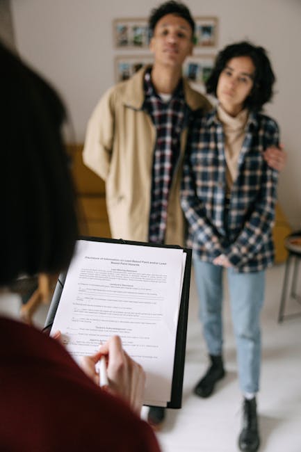 A photograph capturing a scene in an indoor setting with neutral lighting. In the foreground, a person wearing a red shirt holds a black clipboard with a document attached, their hand gripping a pen and appearing to take notes or review the paper. Behind this person, slightly out of focus, are two young adults standing close together; one is a young man with short dark hair, dressed in a beige jacket over a checkered shirt, and the other is a young woman with shoulder-length dark hair, wearing a blue and white plaid shirt. They are positioned on a light-colored floor in front of a white wall decorated with small framed photographs or artwork. The scene appears professional or casual, with the individuals engaged in consultation or discussion, possibly related to property or service agreements, aligning with themes of independent collection or private waste handling, as suggested by the context of rubbish removal services.